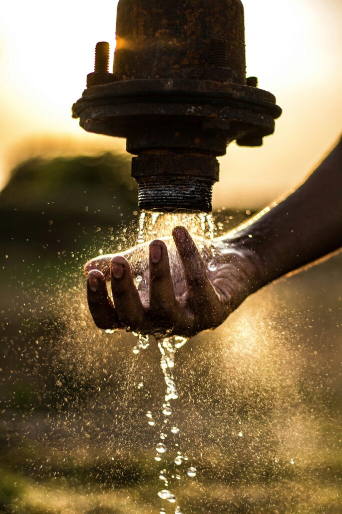 A person washes their hand under a rustic water pipe, capturing the essence of fresh water at sunset.